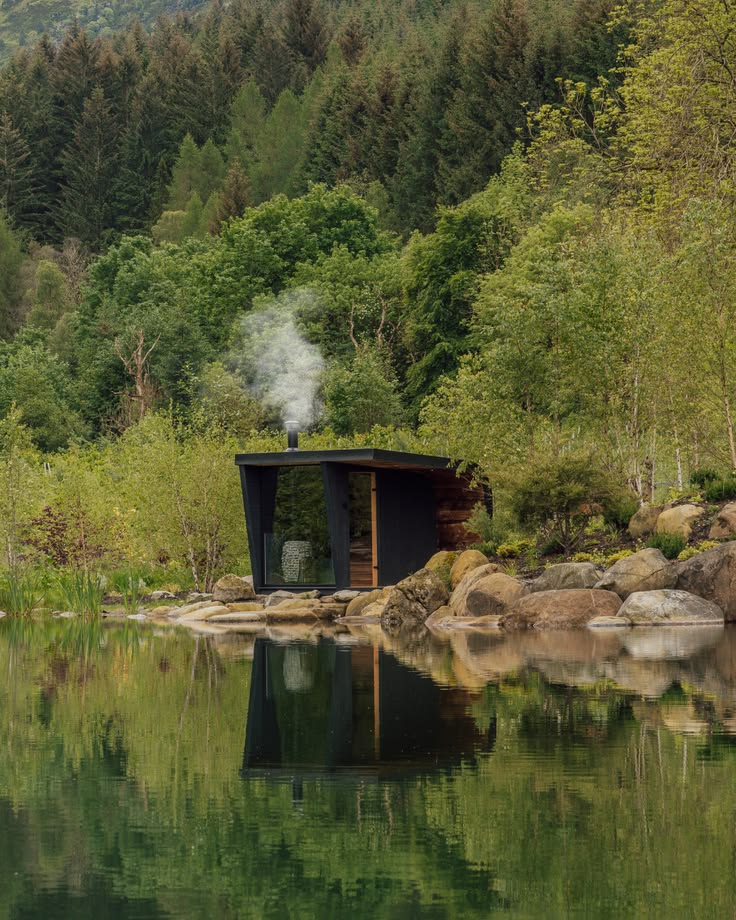 Small dark sauna cabin on rocks at the edge of a green lake, smoke from the chimney