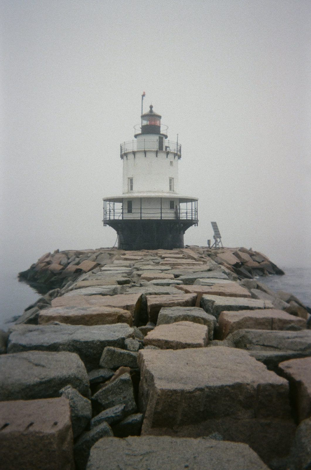 A small lighthouse at the end of a granite breakwater in fog