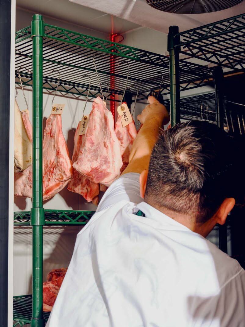 Pork sides hanging from rails in a cold room, a butcher reaching up to a hook
